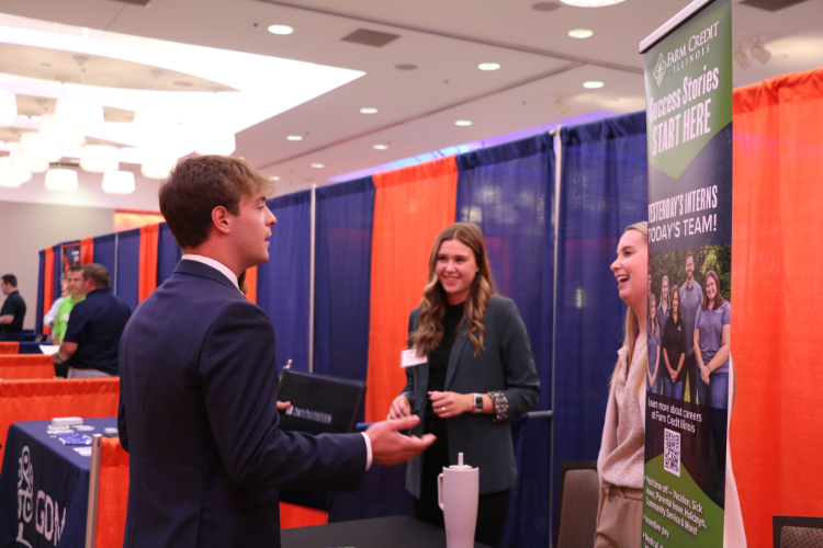 a male in business professional attire speaking to two female employers at a company booth in career fair setting